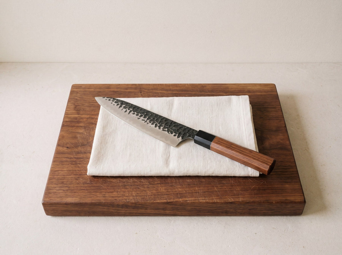 Chef knife with a hammered blade and rosewood handle on a folded linen towel on a walnut cutting board