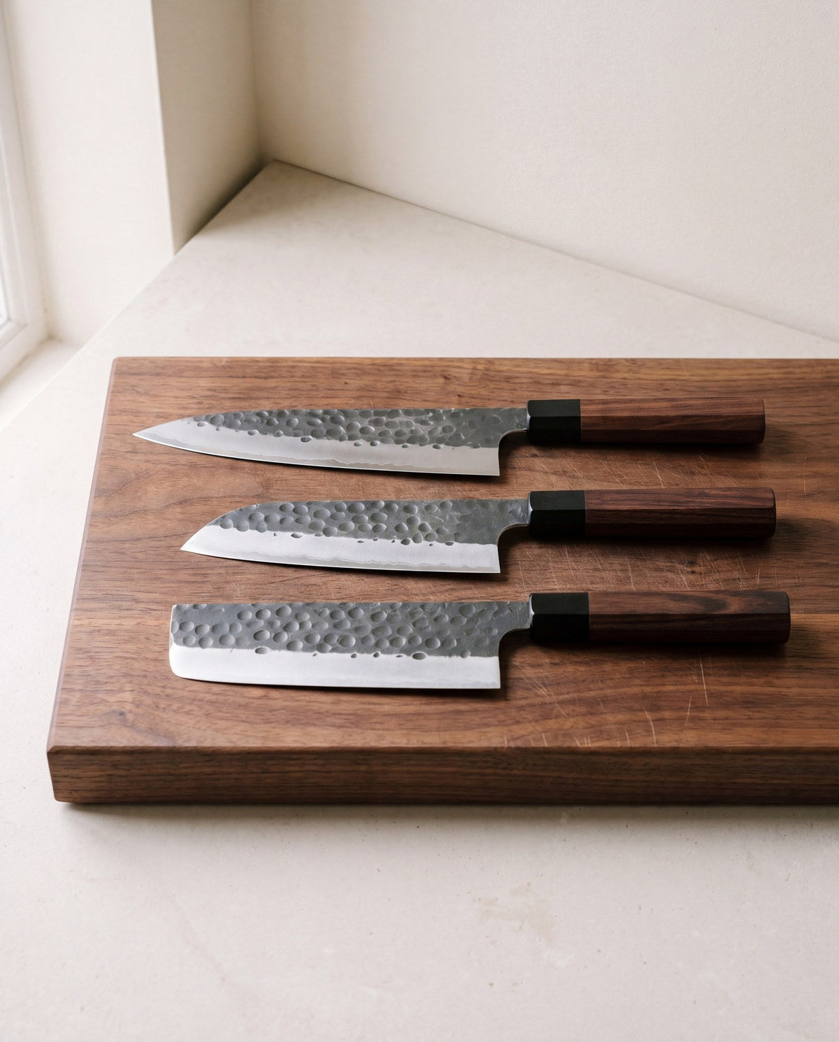 Gyuto, Santoku and Nakiri knives with hammered blades and octagonal wooden handles on a walnut cutting board